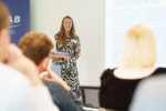 A female speaker talking about her presentation which is displayed large on the wall to a room full of guests. They are in the NIAB building, Cambridge.