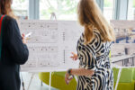 Two women together, one is pointing to an A1 display board on an aisle featuring floorplans and a table of information. They are in the foyer of the NIAB building, Cambridge.