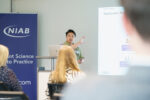 A male speaker talking about his presentation which is displayed large on the wall to a room full of guests. They are in the NIAB building, Cambridge.