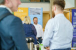 Guests talking and smiling in a bright open space while standing in front of information banners in the foyer of the NIAB building in Cambridge, UK.